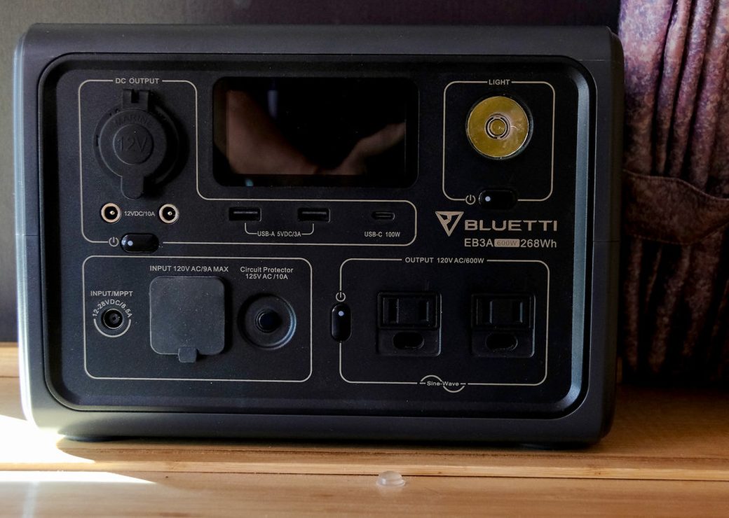 A dark grey portable power station seen from the front: display, a 12 V socket, USB ports and two AC outlets