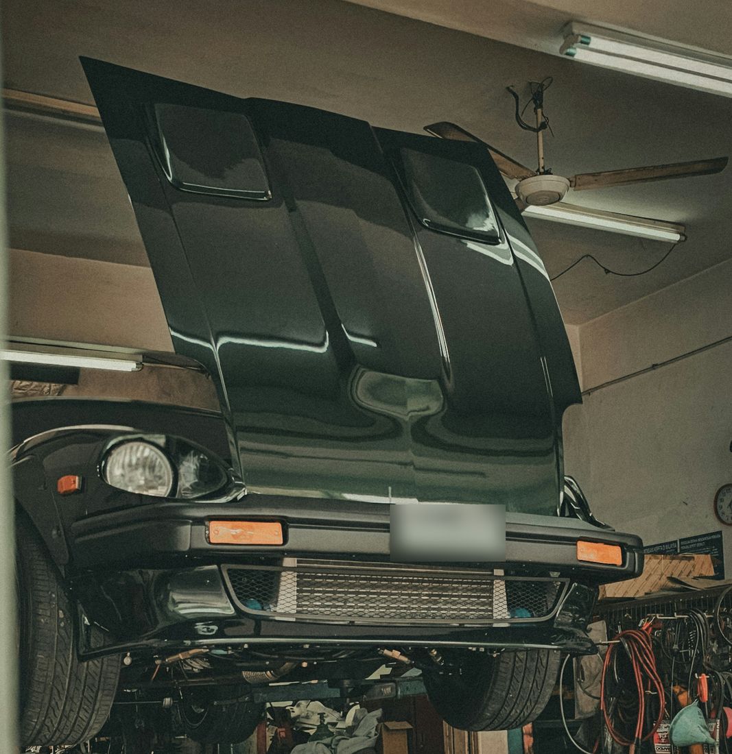 A dark coupe raised on a two-post lift, hood open, in a busy independent workshop