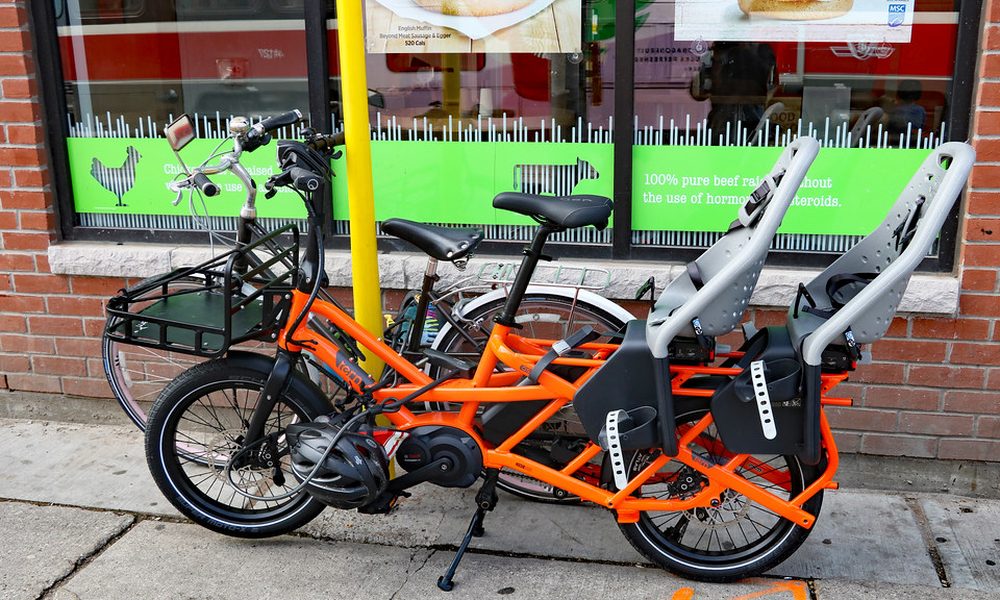 An orange longtail cargo e-bike with two child seats, parked on a city pavement