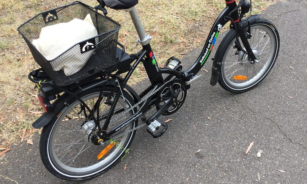 A black folding e-bike with a front basket on a park path