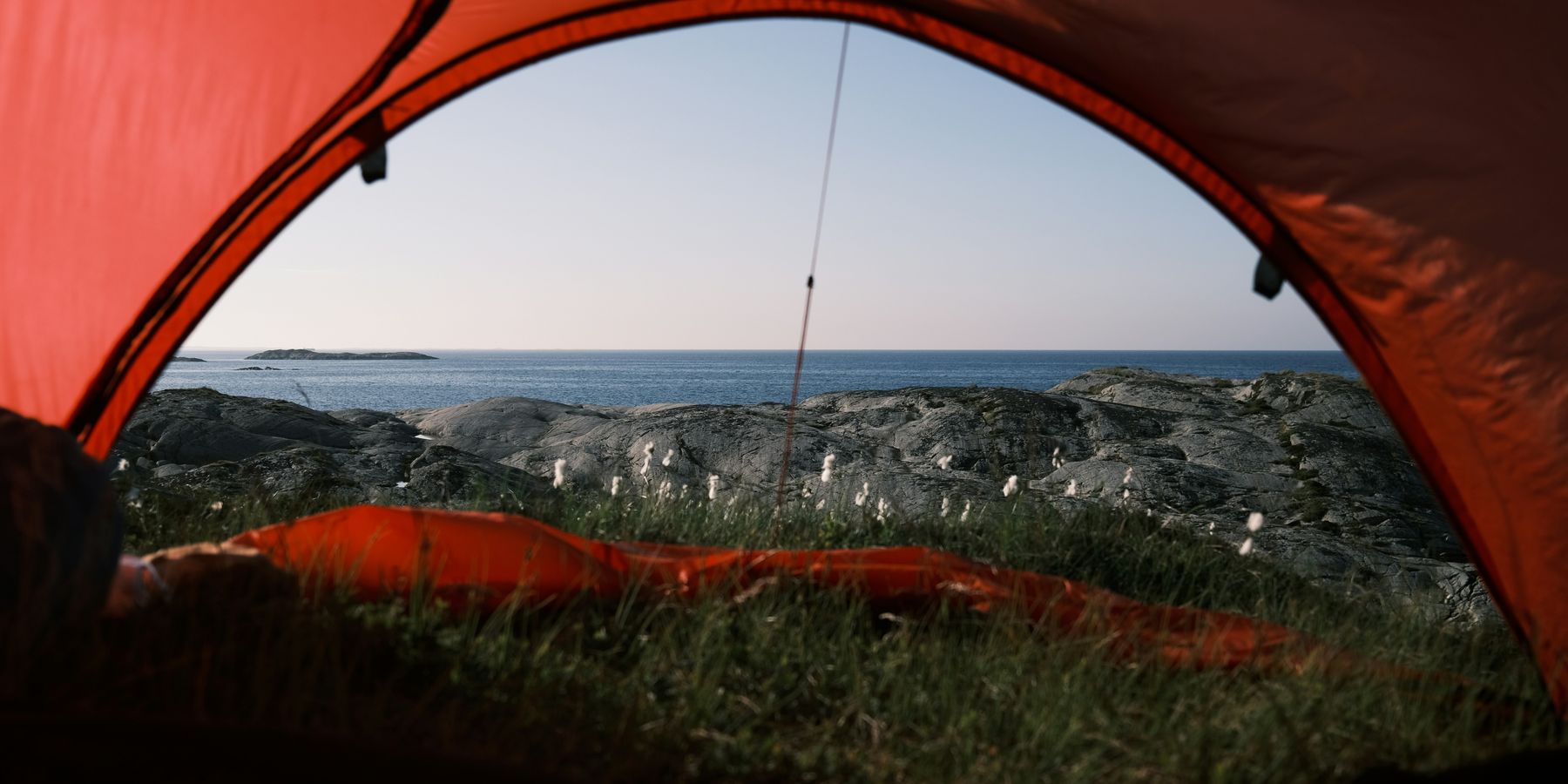 Looking out through the open door of an orange tent at granite slabs, cotton grass and a calm sea