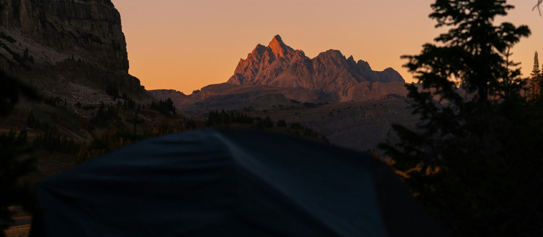 A tent in the foreground at dusk, a jagged peak lit orange by the last sun beyond it