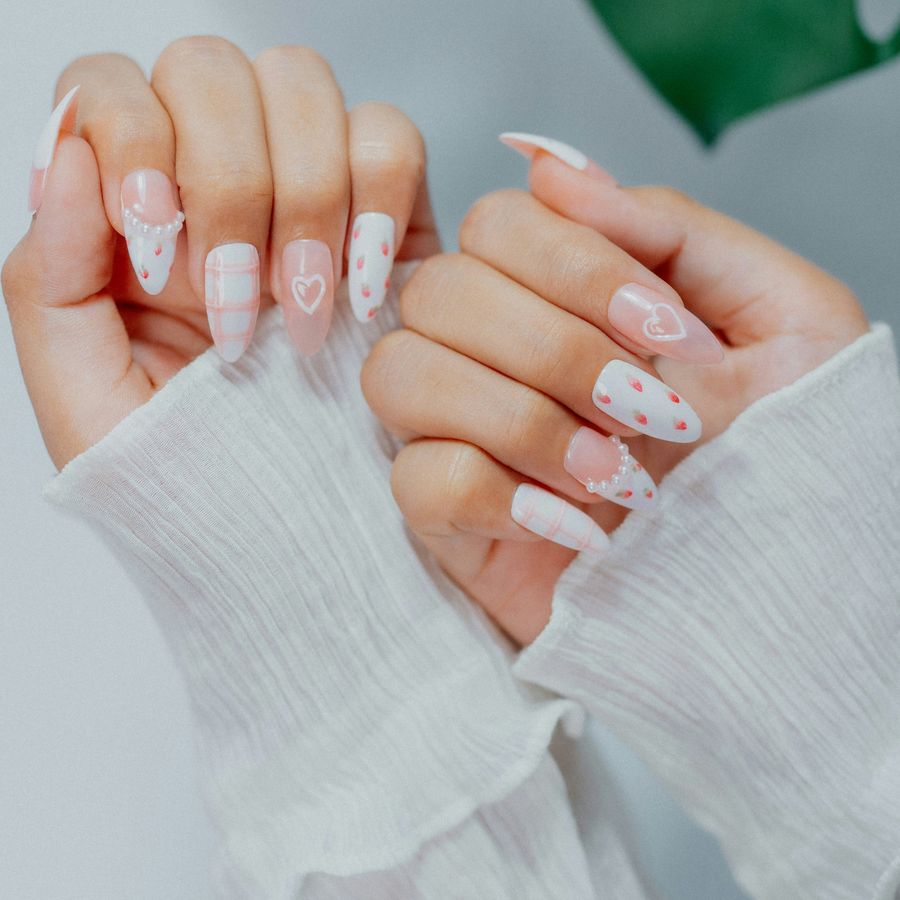 Two hands wearing almond press-on nails painted with strawberries, hearts, gingham and pearls